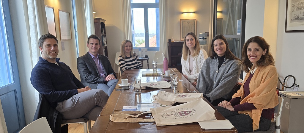 Six people are sitting around a wooden table in a well-lit room. They are facing the camera and appear to be involved in a meeting or discussion. The table has a few items on it, including coffee cups, a water bottle, and a couple of fabric bags.