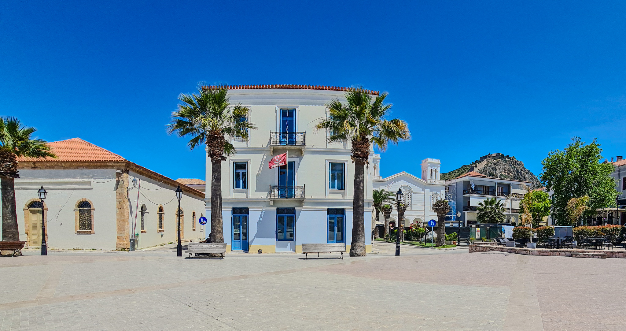 A sunlit town square featuring a cream-colored three-story building with blue window frames and a balcony, flanked by palm trees and benches. To the left is an older building with arched windows and a tiled roof. In the background, there's a white church, residential buildings, more palm trees, parked cars, and a hill with castle ruins on top under a clear blue sky.