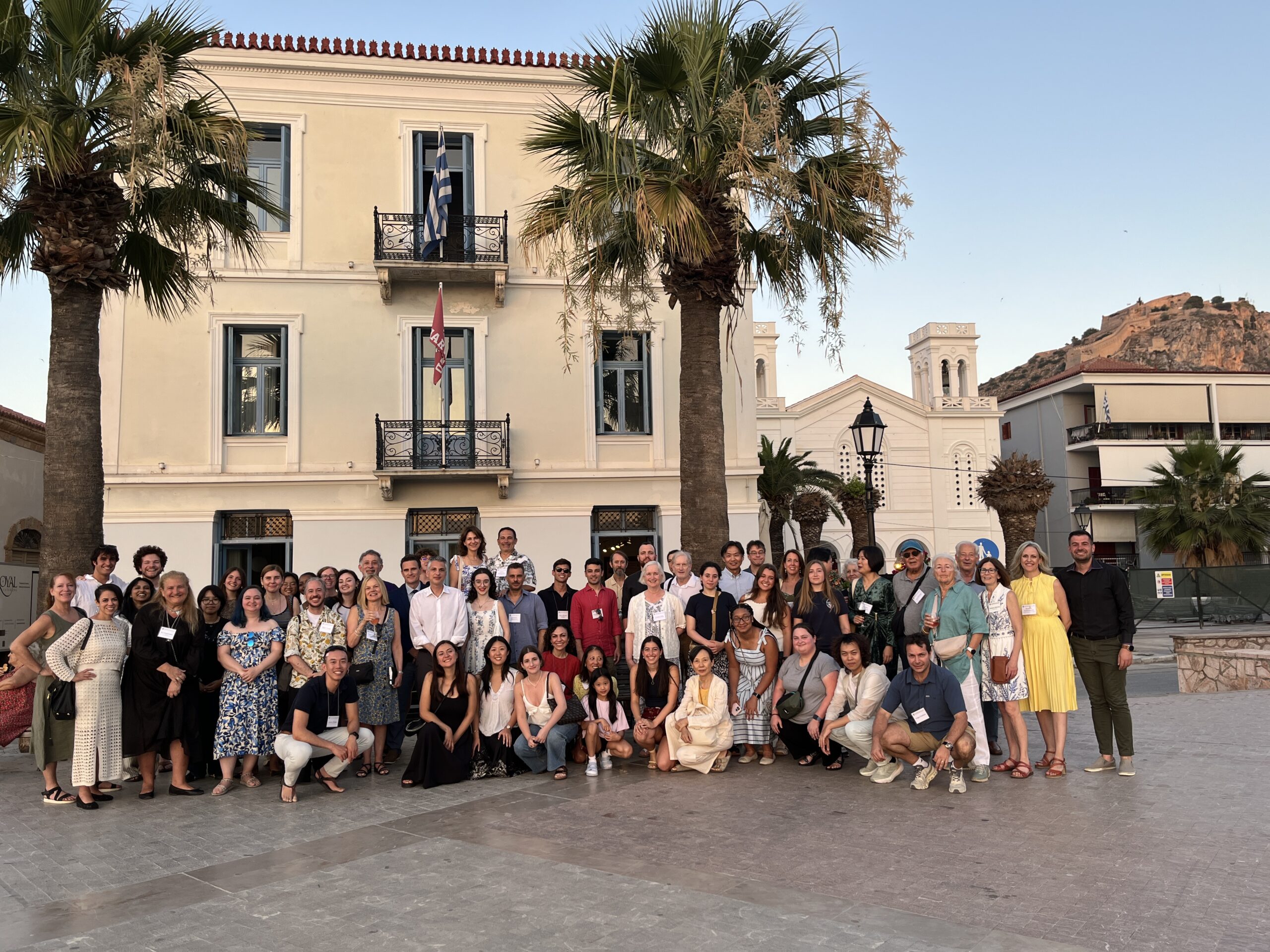A large, diverse mix of men and women pose together for a group photo outdoors in front of a neoclassical building with balconies and flags.