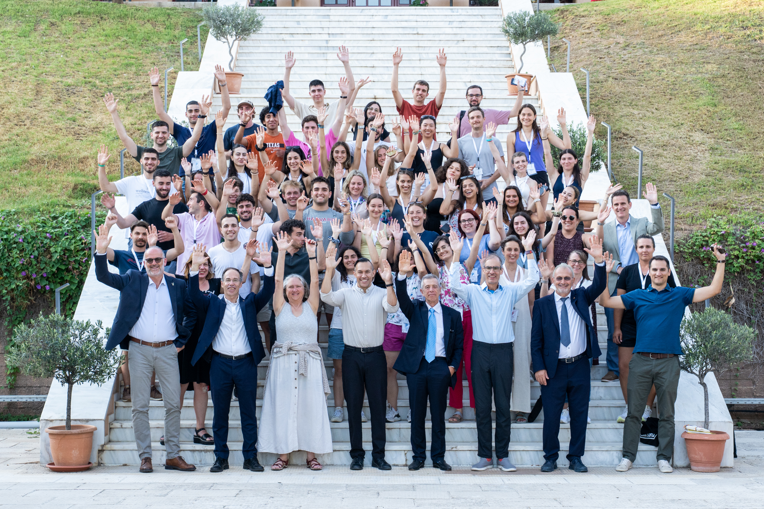 A large group of people pose together on outdoor stairs, smiling and raising their hands in the air for a group photo.