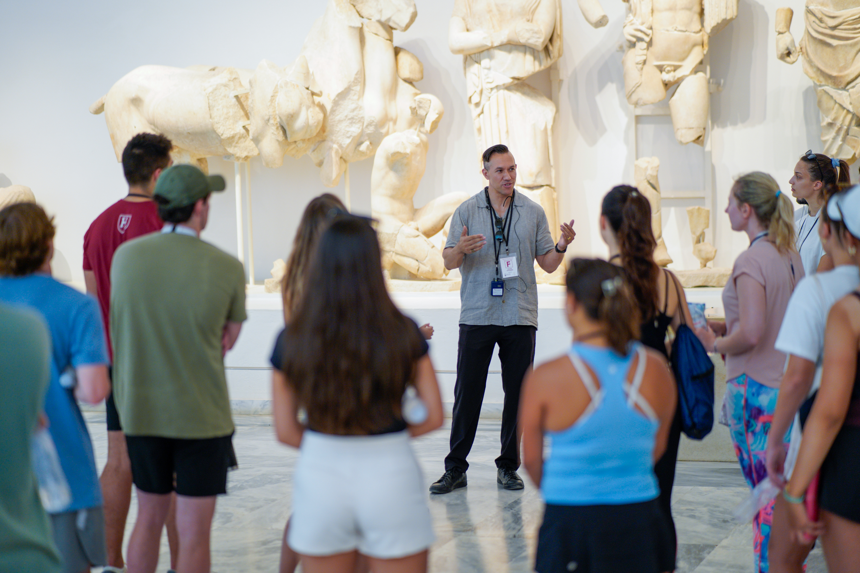 Guided by a faculty member gesturing as he speaks, a group of young men and women listens attentively. Behind him are ancient marble sculptures and fragments displayed on a wall.