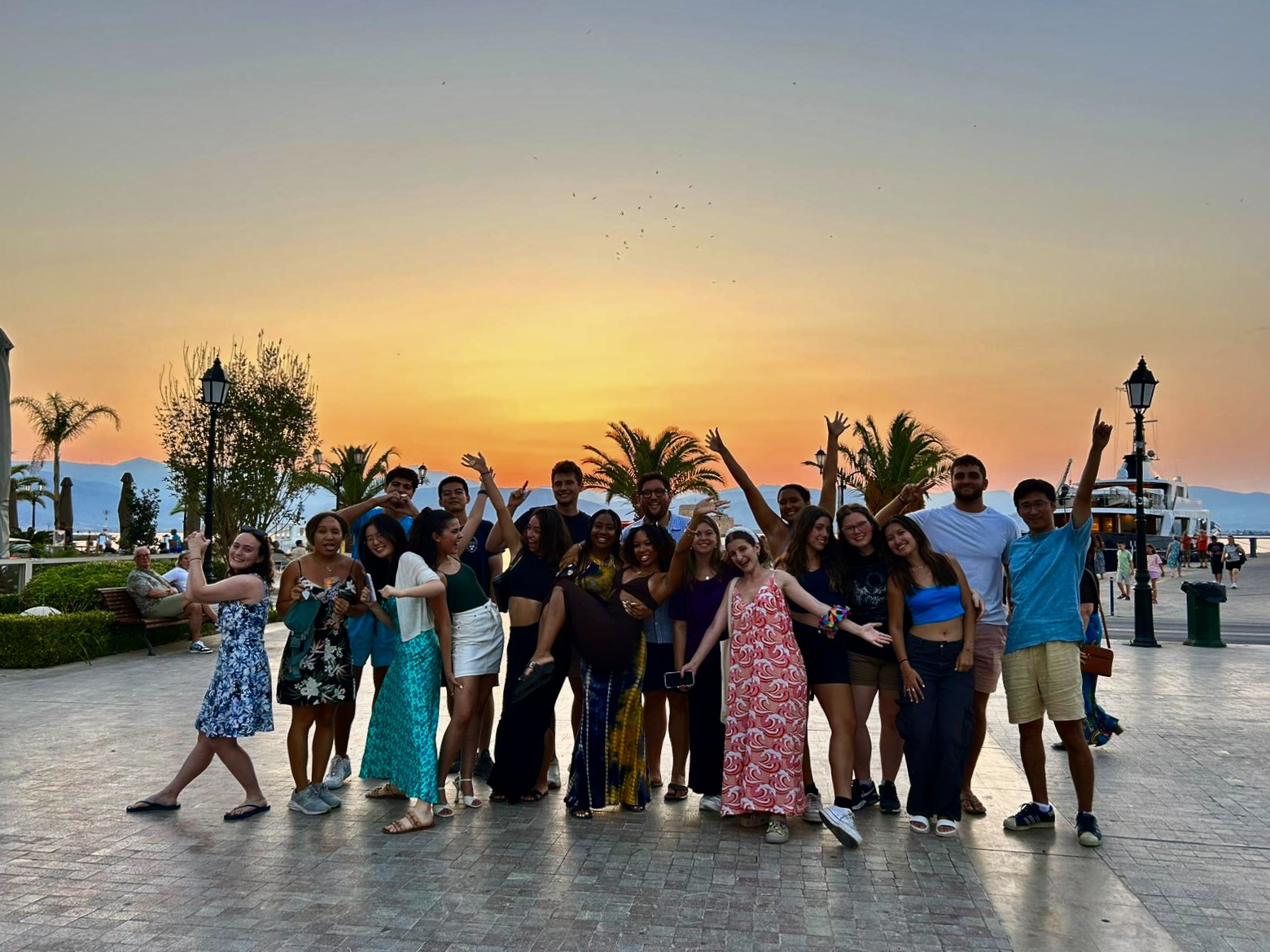 Large group of young adults posing together at sunset on a waterfront promenade.