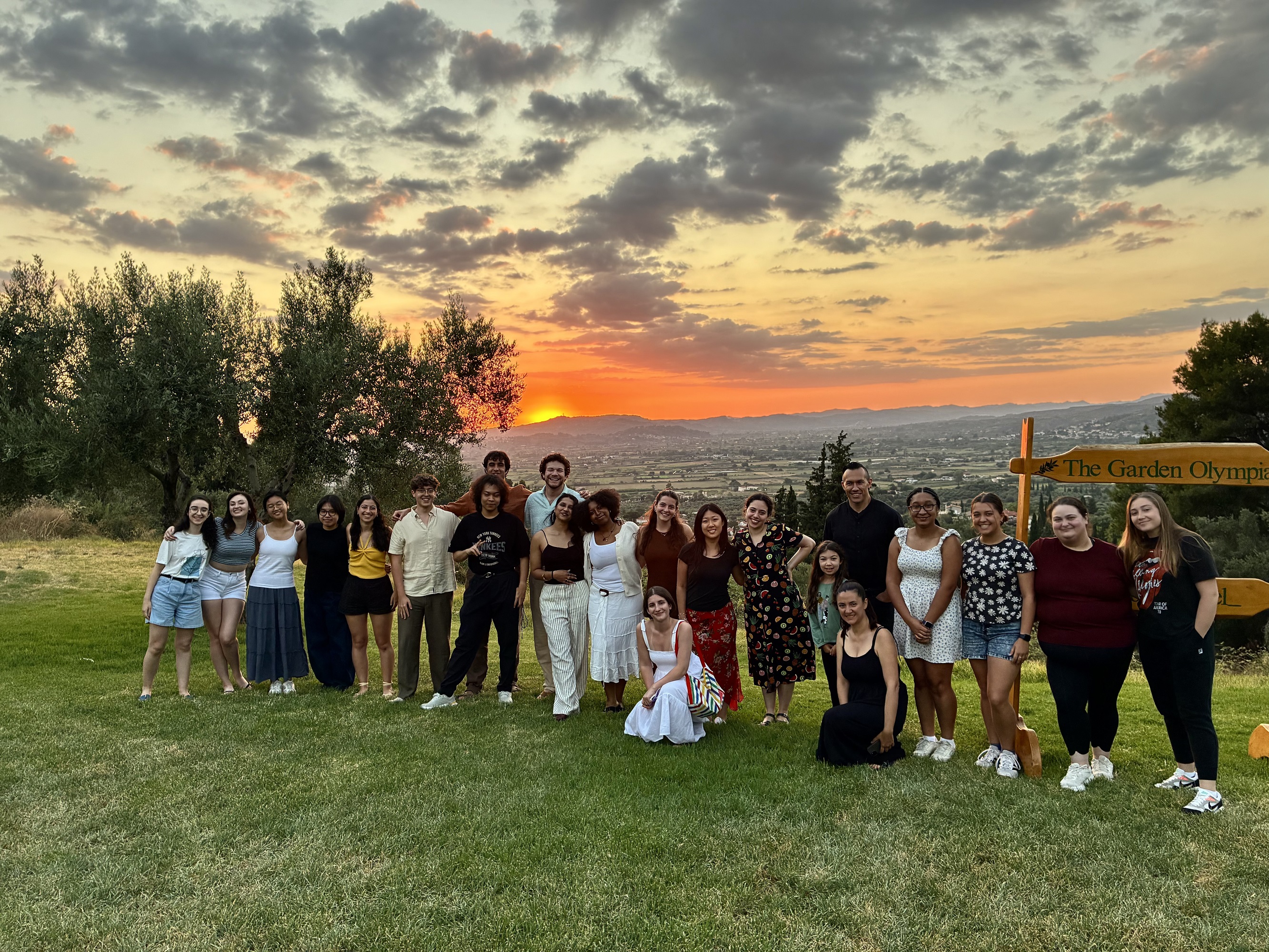 Group of people standing and sitting on grass with sunset and mountains behind them.