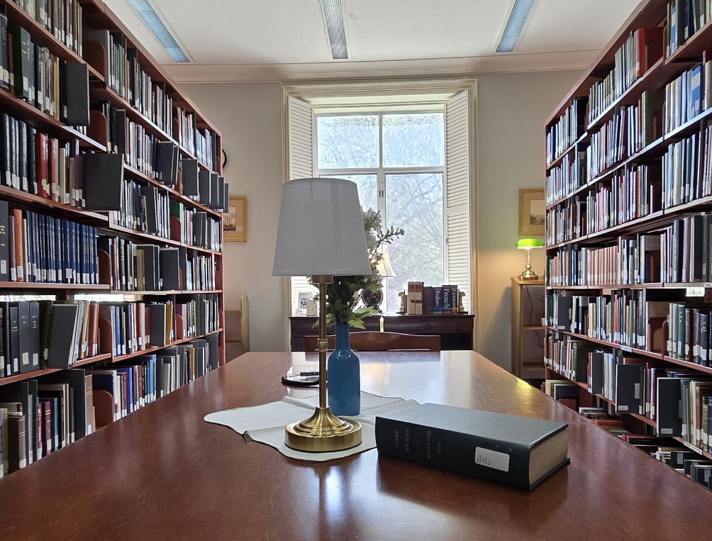A quiet library room with bookshelves lining the walls, a large wooden table, a lamp, and a book.