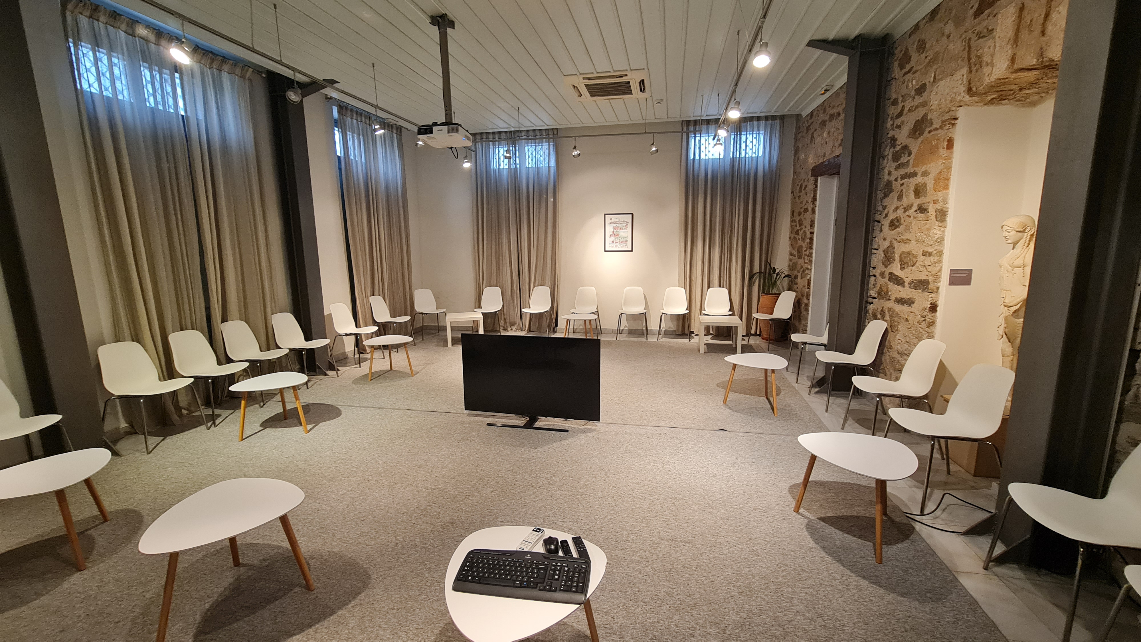 Conference room with chairs arranged in a circle, small white tables, a large screen in the center, and one wall made of exposed stone.