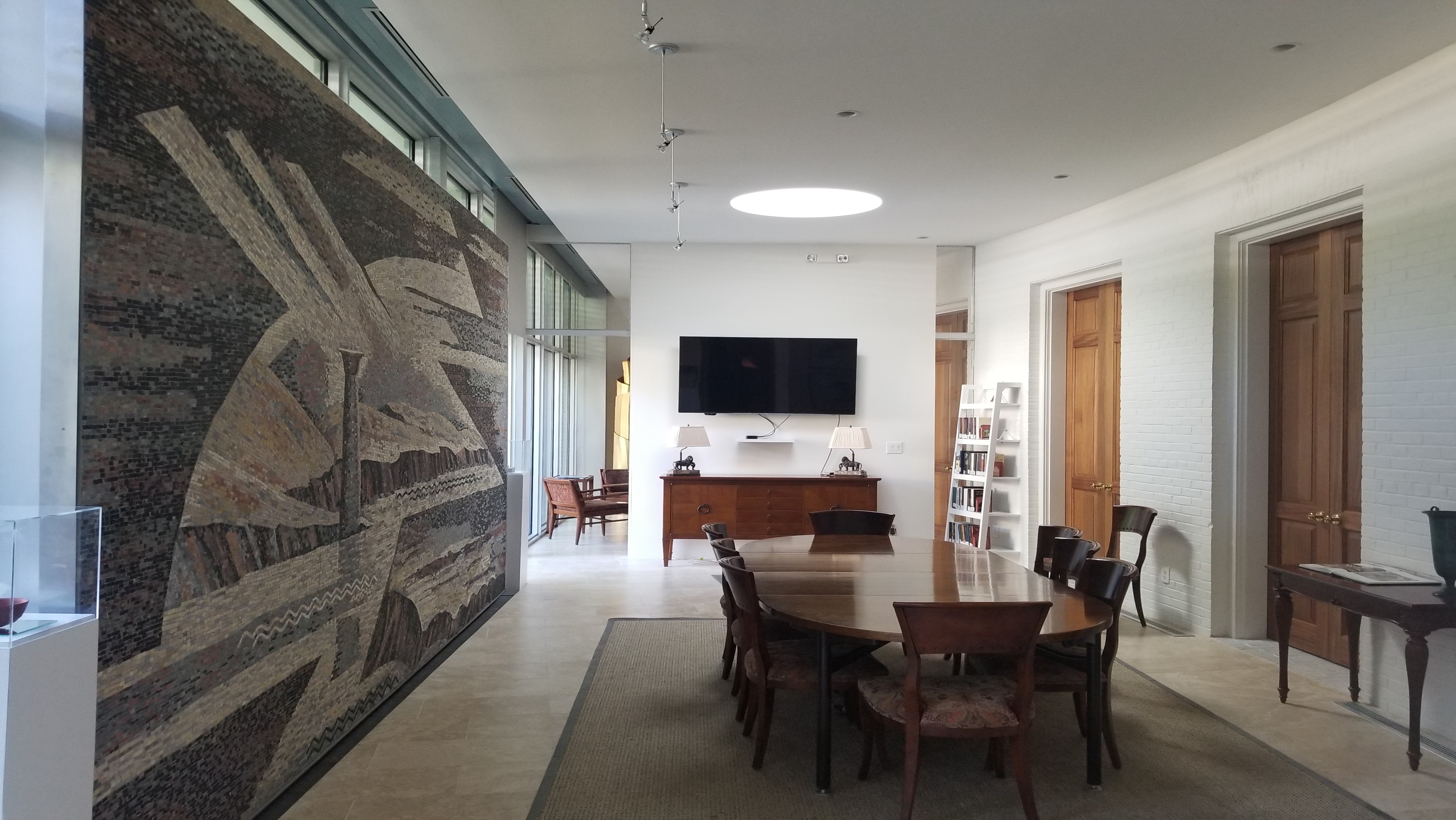 Long, bright meeting room with a large mosaic wall, oval wooden conference table and chairs, screen on the far wall, and bookshelves by wooden doors.