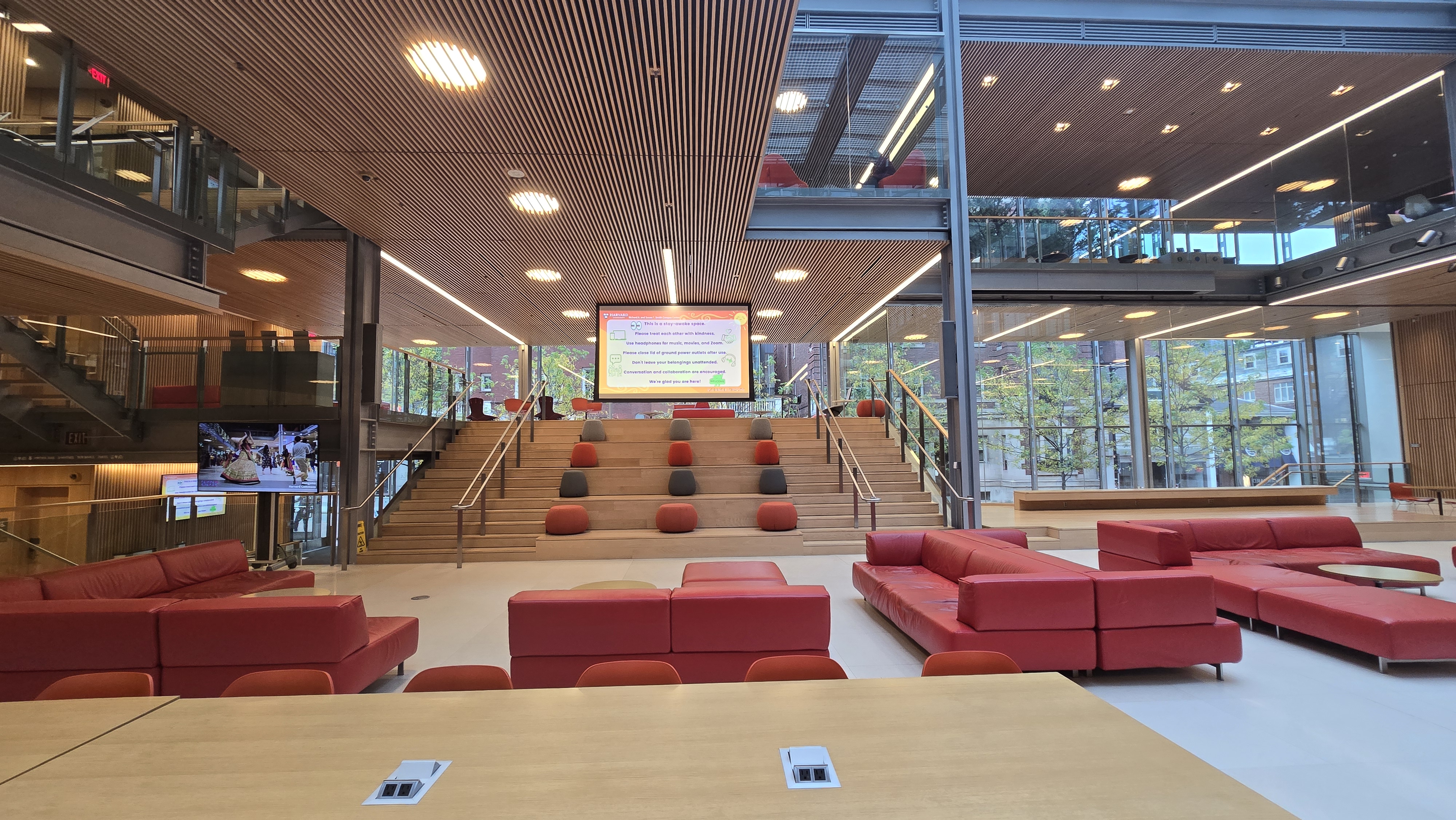 Modern, open lobby with floor‑to‑ceiling windows, red couches, and wide wooden steps with cushions leading up to a large screen.