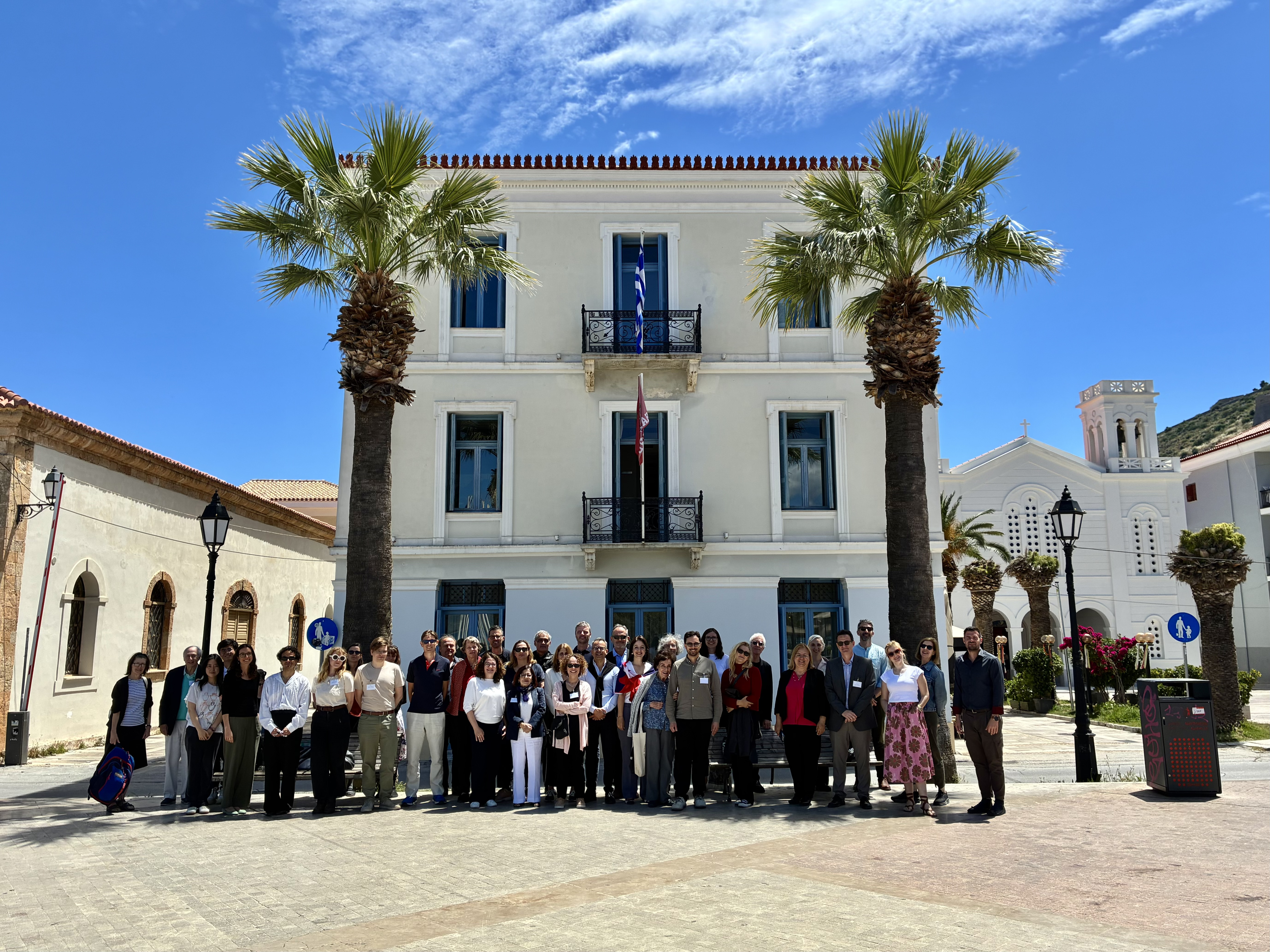 Group photo of conference participants standing in front of a white neoclassical building with palm trees.