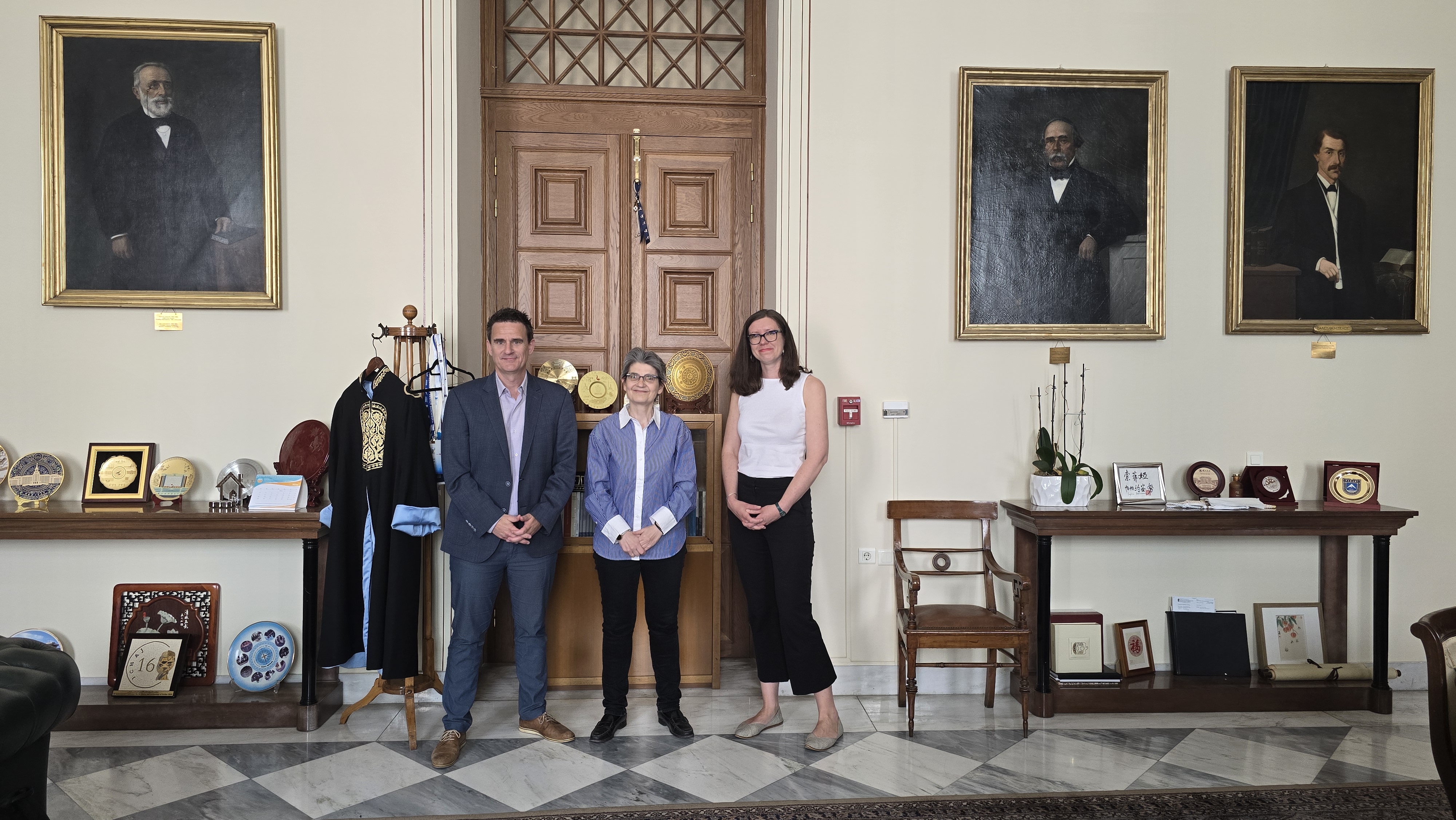 Three people standing in a formal office with a large wooden door and framed portraits.