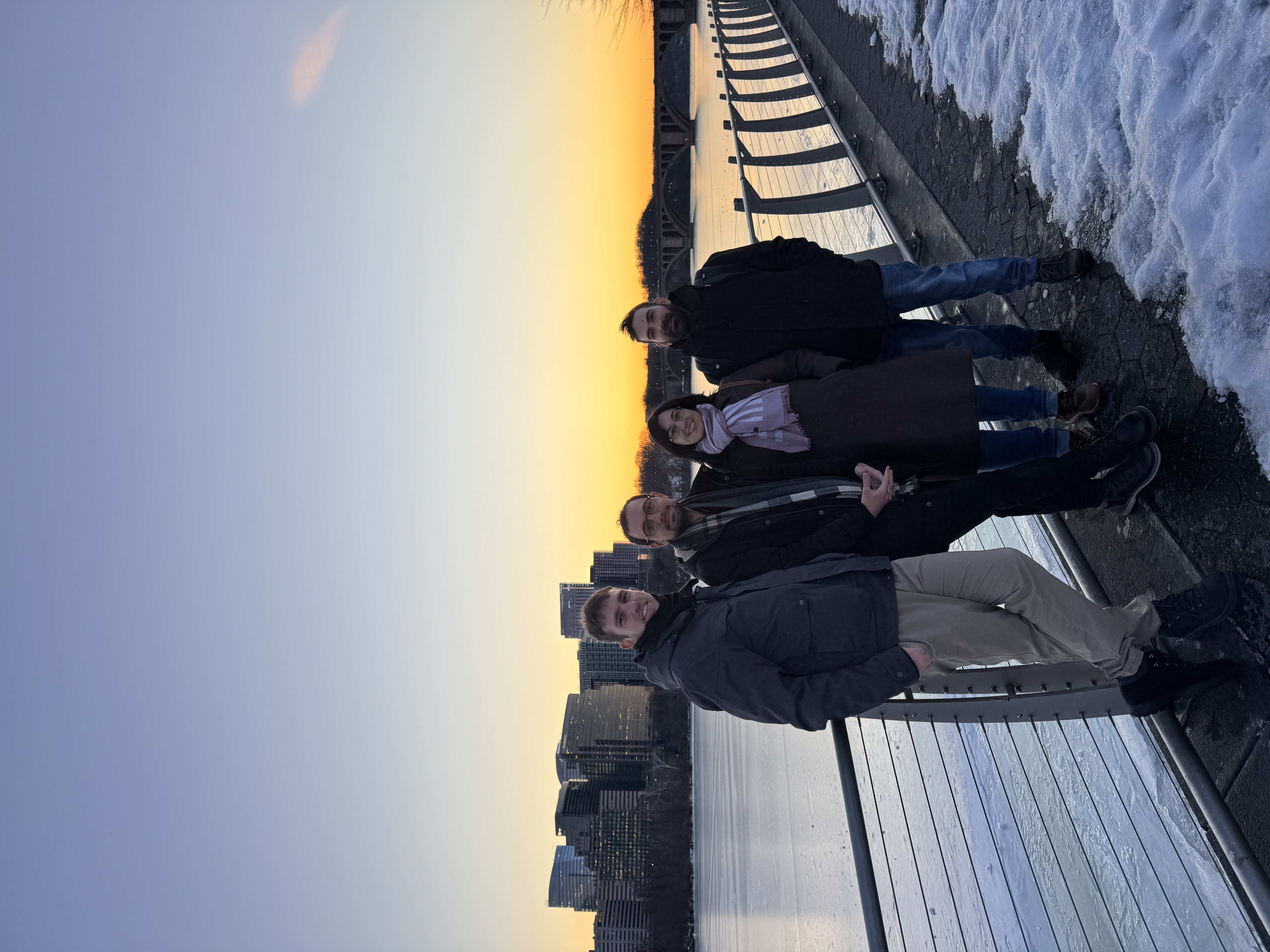 Four people standing by a riverside railing at sunset in winter