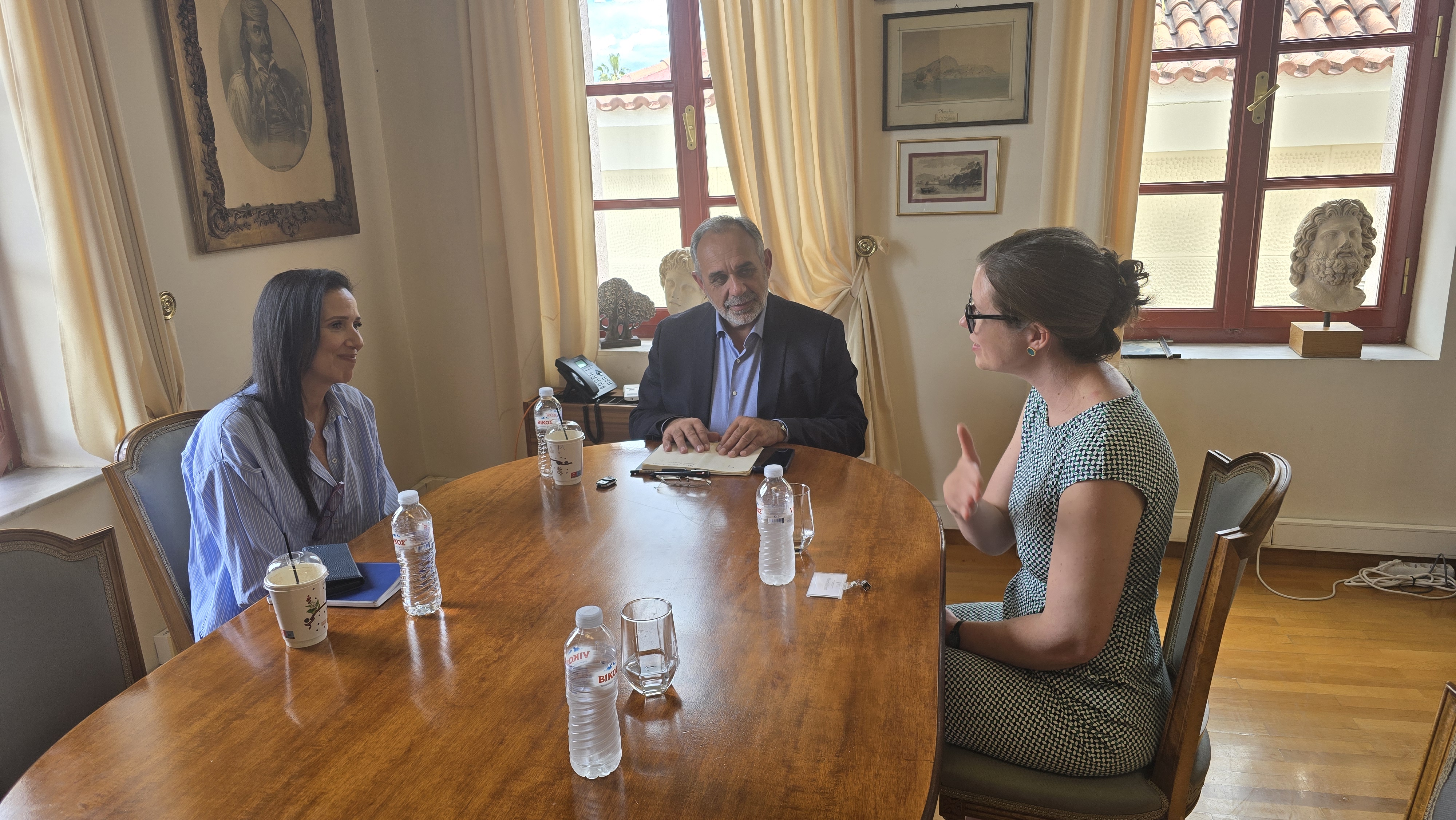Three people seated around a wooden table in a sunlit office, talking.