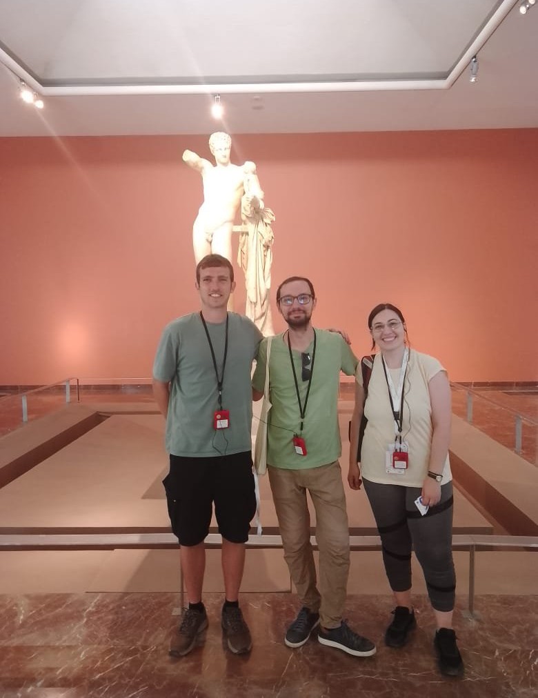 Three visitors wearing casual clothes and museum badges pose in front of a large classical white statue in a gallery with reddish walls.