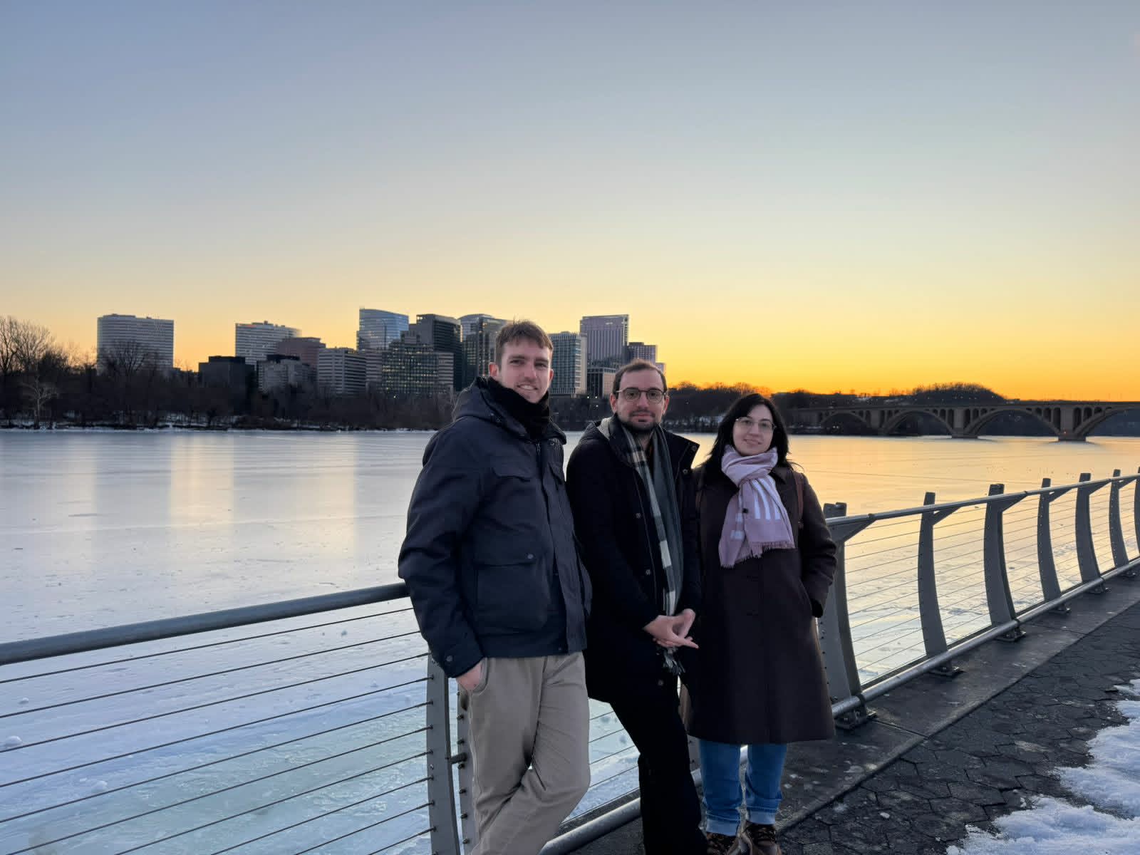 Three people standing by a riverside railing at sunset in winter.