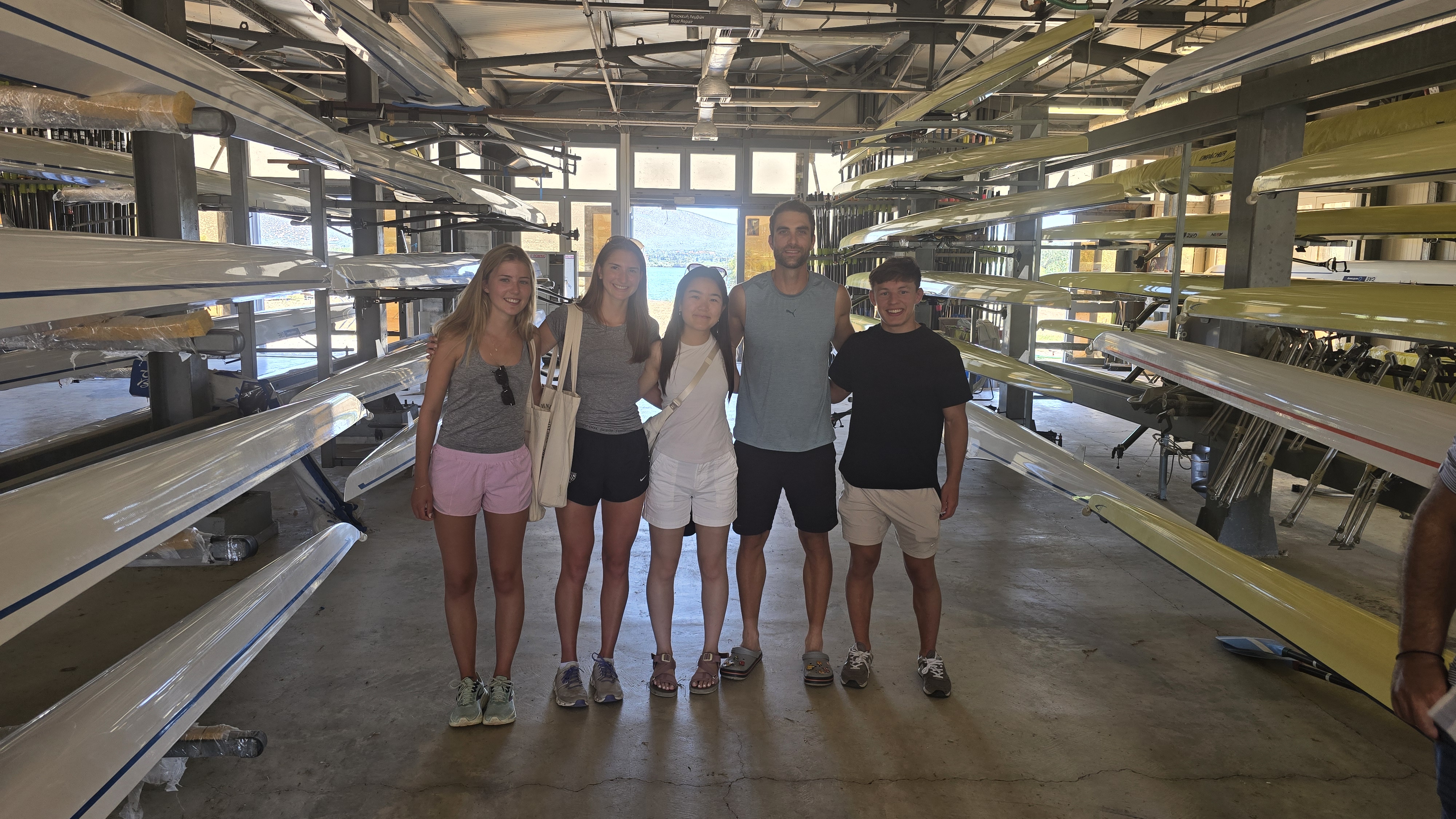 Harvard student-athletes along Stefanos Ntouskos stand inside the Schinias Olympic Rowing and Canoeing Centre boathouse, surrounded by rowing shells. 