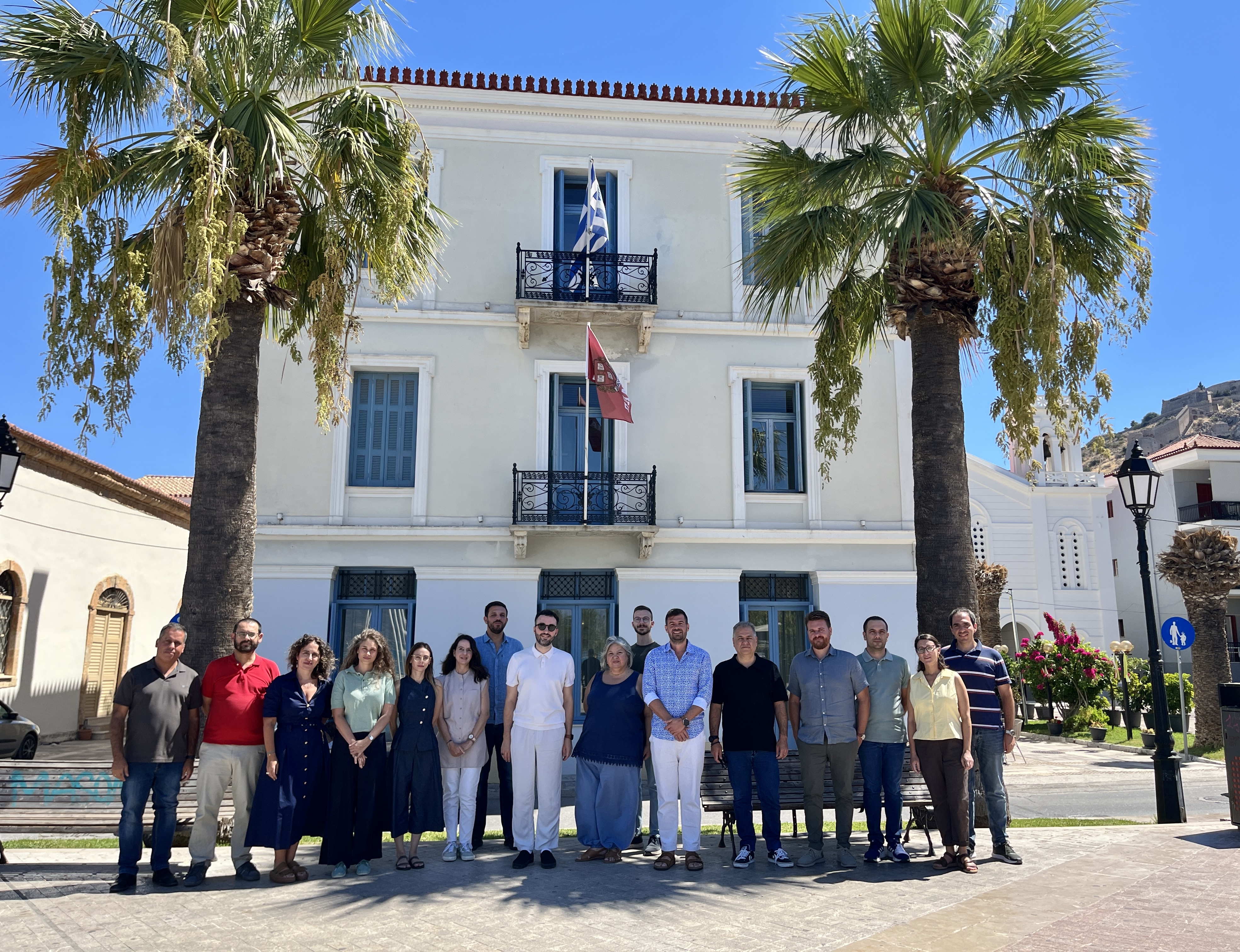 Group portrait of the workshop participants outside the CHS Greece building in Nafplio, between two palm trees.