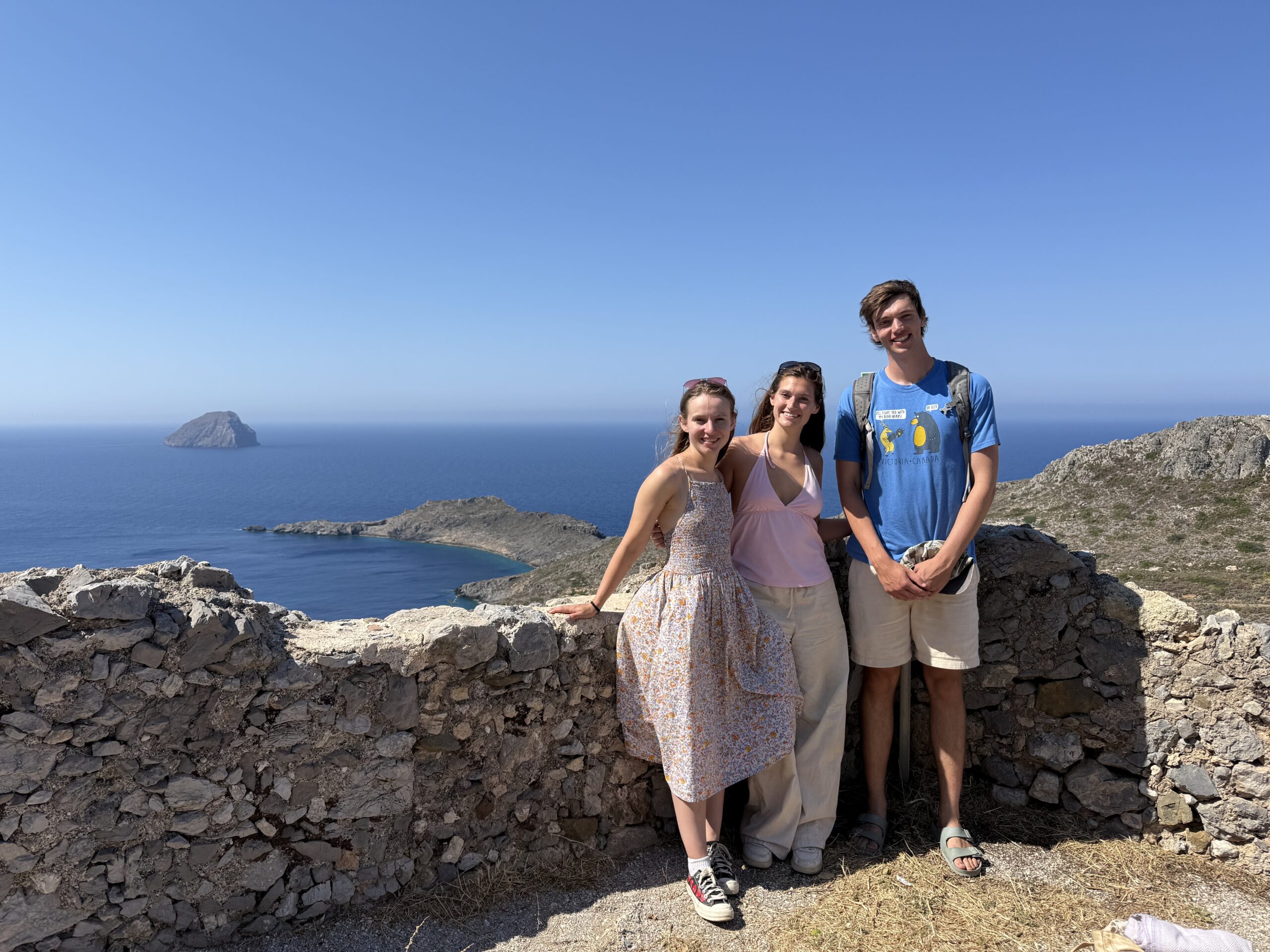 Three participants posing at a scenic coastal viewpoint, with rocky islands and blue sea in the background.