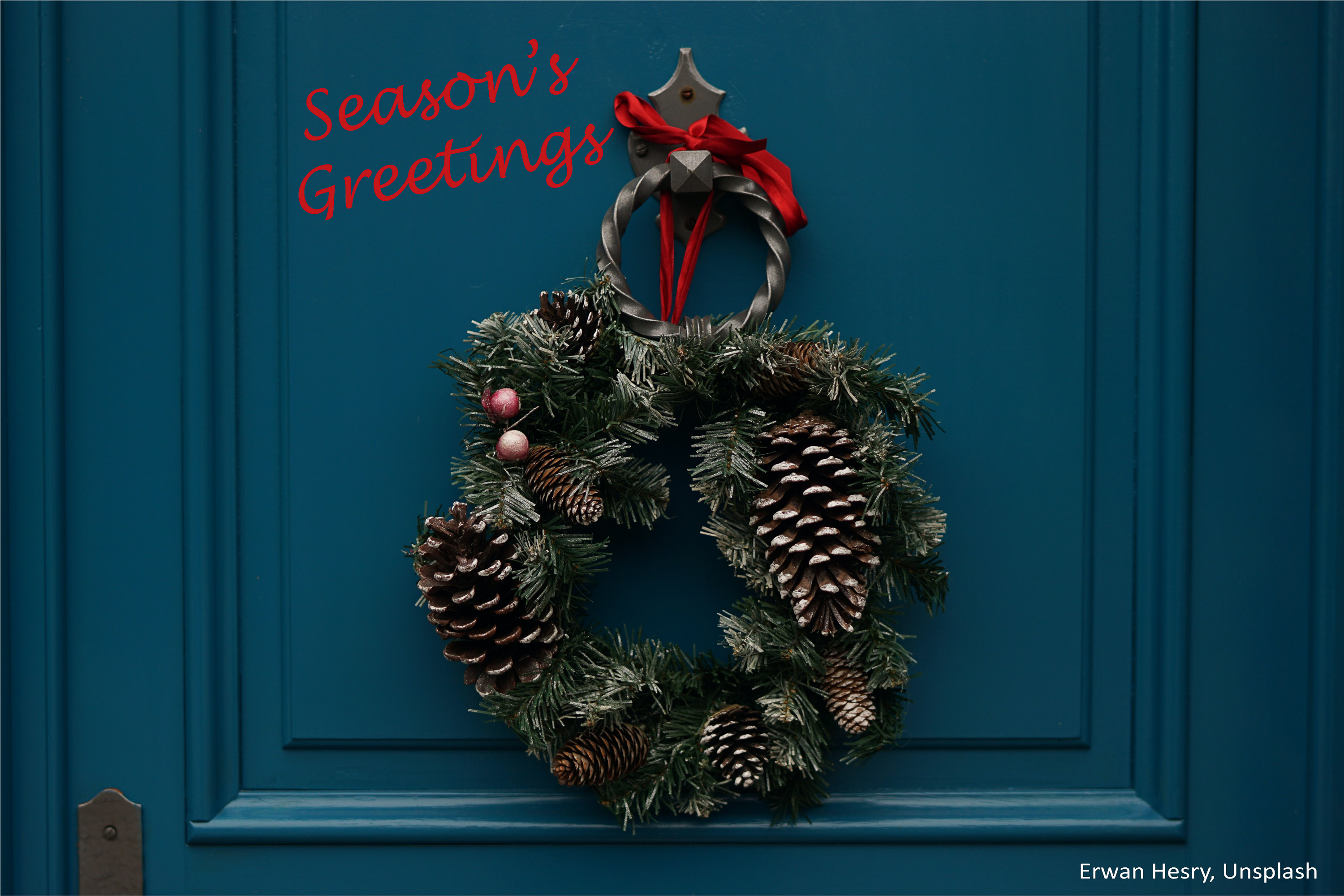 Christmas wreath with pinecones hanging from a red ribbon on a knocker of a blue door. “Season’s Greetings” message is also added, and photo credit to Erwan Hesry, Unsplash.