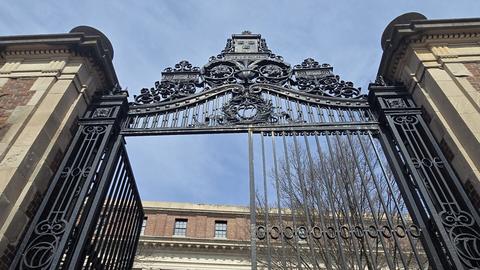 A low-angle view of a grand wrought-iron gate, open and ornately decorated at the top, leading to a large brick building with tall windows.