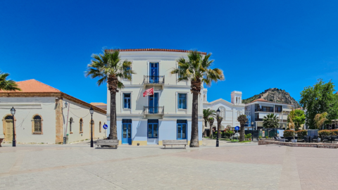 A sunlit town square featuring a cream-colored three-story building with blue window frames and a balcony, flanked by palm trees and benches. To the left is an older building with arched windows and a tiled roof. In the background, there's a white church, residential buildings, more palm trees, parked cars, and a hill with castle ruins on top under a clear blue sky.