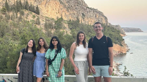 Five people stand side by side, smiling for the camera, with a scenic backdrop of rocky cliffs, green trees, and the sea at sunset. The group is casually dressed and appears relaxed, enjoying the outdoor setting.