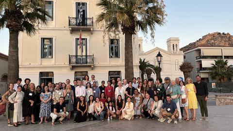 A large, diverse mix of men and women pose together for a group photo outdoors in front of a neoclassical building with balconies and flags.