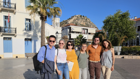 Six adults smile and pose together in a sunny town square. Behind them are a palm tree, pastel buildings, and a rocky hill with a fortress on top, all under a clear blue sky.