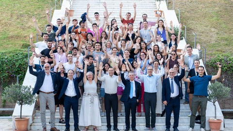 A large group of people pose together on outdoor stairs, smiling and raising their hands in the air for a group photo.