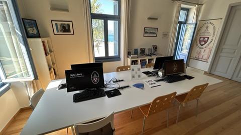 Bright study room with computers on a white table, wooden chairs, bookshelves, and large windows letting in sunlight.