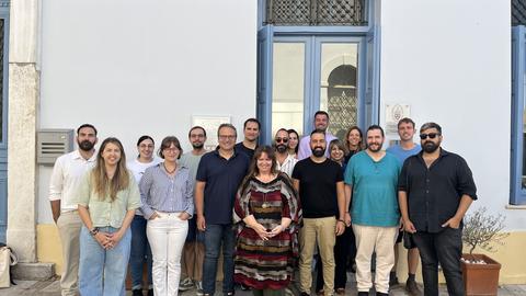 A group of about 20 people pose for a group photo on a sunny day outside of a neoclassical building.