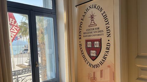 A banner for the Center for Hellenic Studies in Greece of Harvard University hangs next to a window overlooking a seaside street with palm trees and flags.