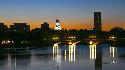 Evening falls over the Charles River, shimmering city lights and the curve of a bridge reflected in tranquil waters.