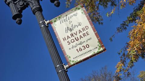A welcoming banner graces a lamppost beneath a blue sky, inviting visitors to the storied Harvard Square.