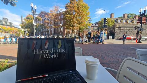 A laptop and a cup of coffee rest on a table in historic Harvard Square, its screen displaying the words "Harvard in the World."