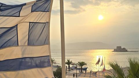Greek flag blowing in the wind above a seaside promenade at sunset, with people walking below and the sun reflecting on the water.