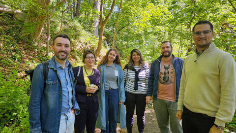 Six people standing and smiling on a sunlit wooded trail.