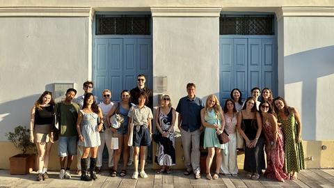 Group photo of program participants and faculty standing outside a neoclassical building with blue doors in warm evening light.