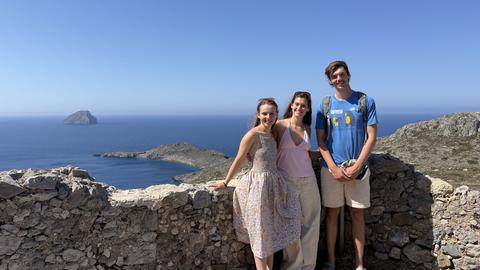 Three participants posing at a scenic coastal viewpoint, with rocky islands and blue sea in the background.