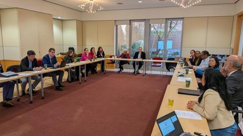 Participants seated around U-shaped tables during a meeting in a conference room with laptops, documents, and large windows.