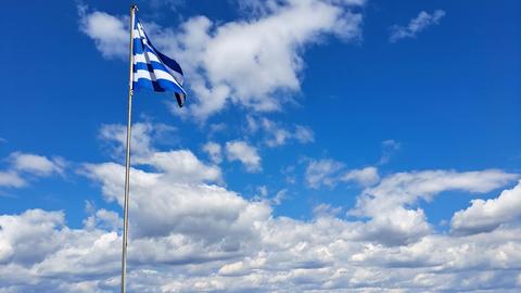 A Greek flag flies on a pole above a stone wall, overlooking the sea and mountains under a bright, partly cloudy sky.