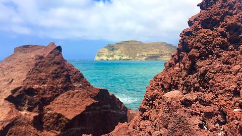 Rocks made by a reddish soil and stone along with the view of the sea and a peninsula in the background..