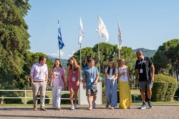 Participants walk together outdoors at the International Olympic Academy in Ancient Olympia, with flags and greenery in the background.