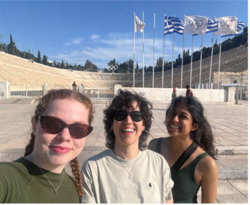 Three participants pose for a photo at the Panathenaic Stadium in Athens, with Olympic flags visible in the background.