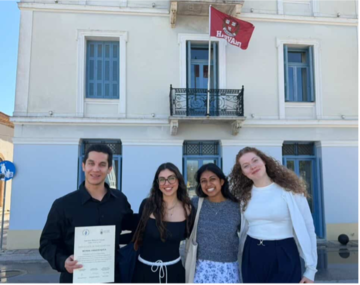 Four participants stand in front of the CHS Greece building in Nafplio beneath a Harvard flag.