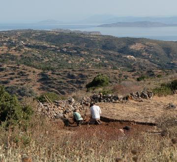 Archaeological excavation overlooking a coastal landscape, with two people working in a dig area on a hillside.