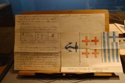 A wooden panel displaying handwritten notes and sketches of preliminary flag designs alongside colorful renditions of the Greek flag.