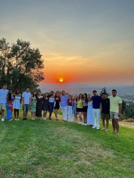 A group of people stands outdoors on a grassy field, posing with their arms around each other. In the background, the sun sets over distant mountains, casting a warm orange and pink glow across the sky, while tall trees and a view of a green valley are framing the sunset.