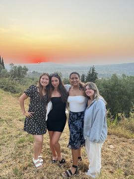 Four young women are posing with their arms around each other on a slope, as the sun sets in the background, casting orange and pink hues across the sky and over the surrounding greenery.