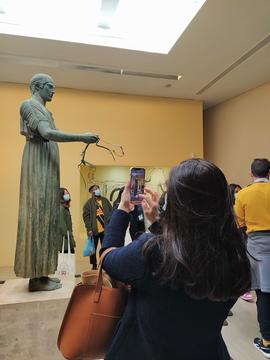 A black-haired woman takes a photo of a bronze statue of a charioteer inside a museum hall, with other visitors visible in the background.
