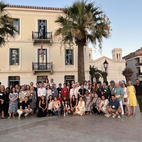 A large, diverse mix of men and women pose together for a group photo outdoors in front of a neoclassical building with balconies and flags.