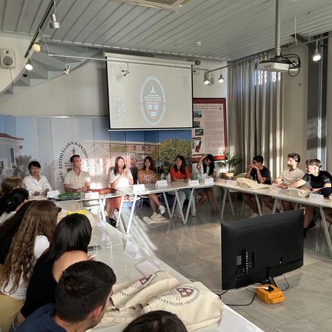 A group of people sit around conference tables in a classroom, engaged in discussion, with a projector screen at the front.
